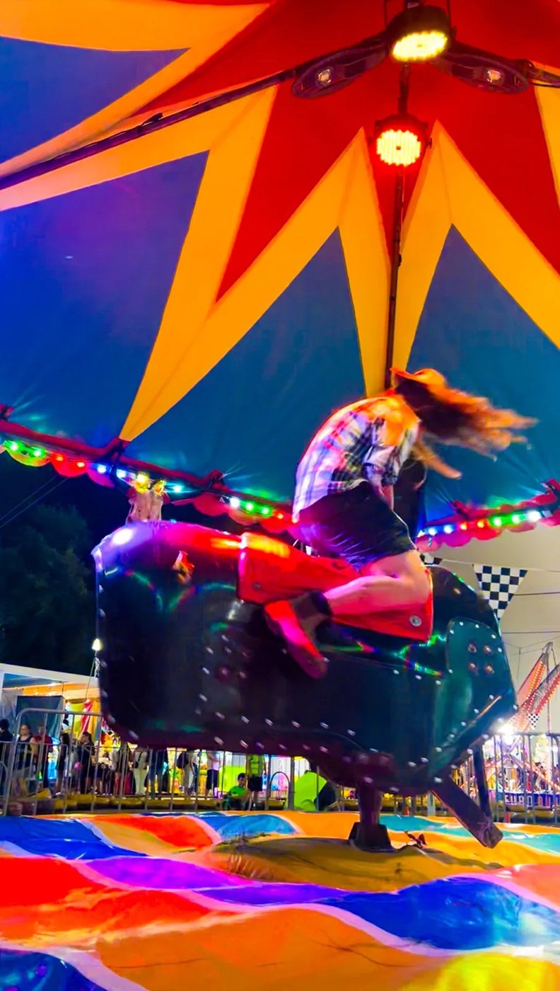 A rider on a mechanical bull under a red, yellow and blue striped tent at night