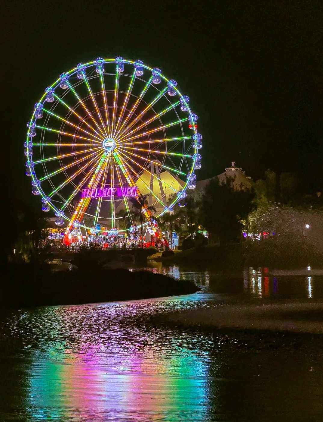 A neon-lit Ferris wheel reflected in the water at night