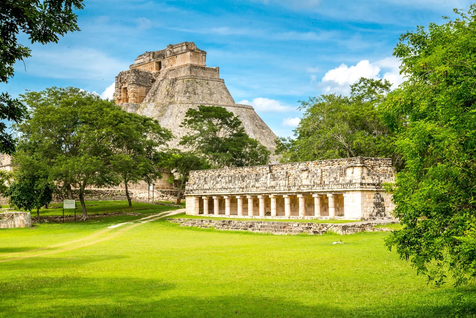 The Pyramid of the Magician rising above trees and a colonnaded Maya building at Uxmal